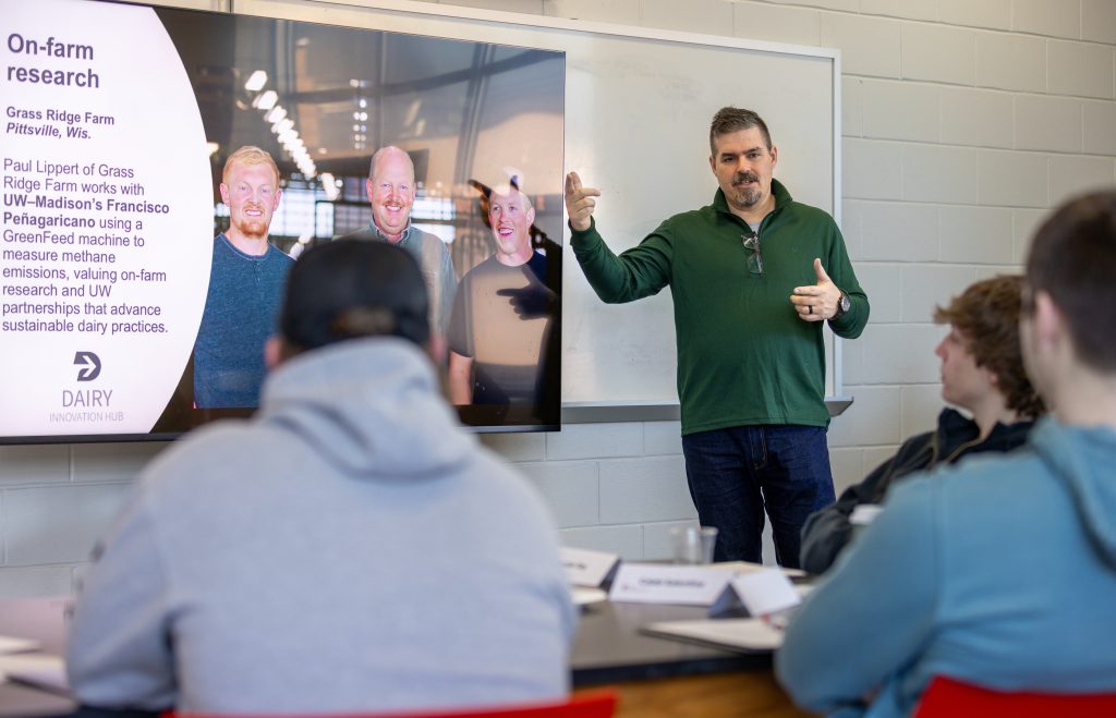 Man speaks in front of monitor to men sitting in audience.