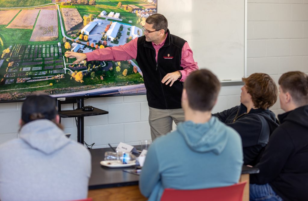 Man speaks in front of monitor to men sitting in audience