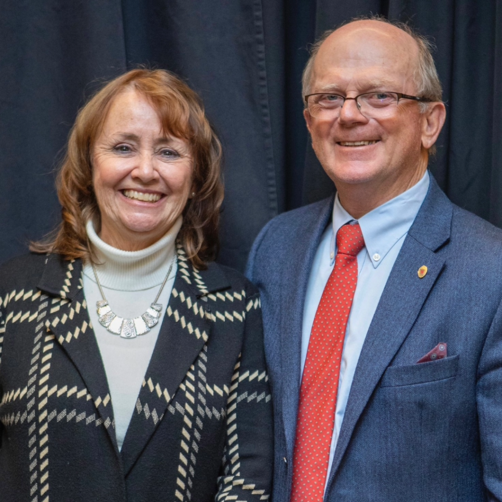 Man with grey hair and woman with red hair stand next to each other looking at camera.